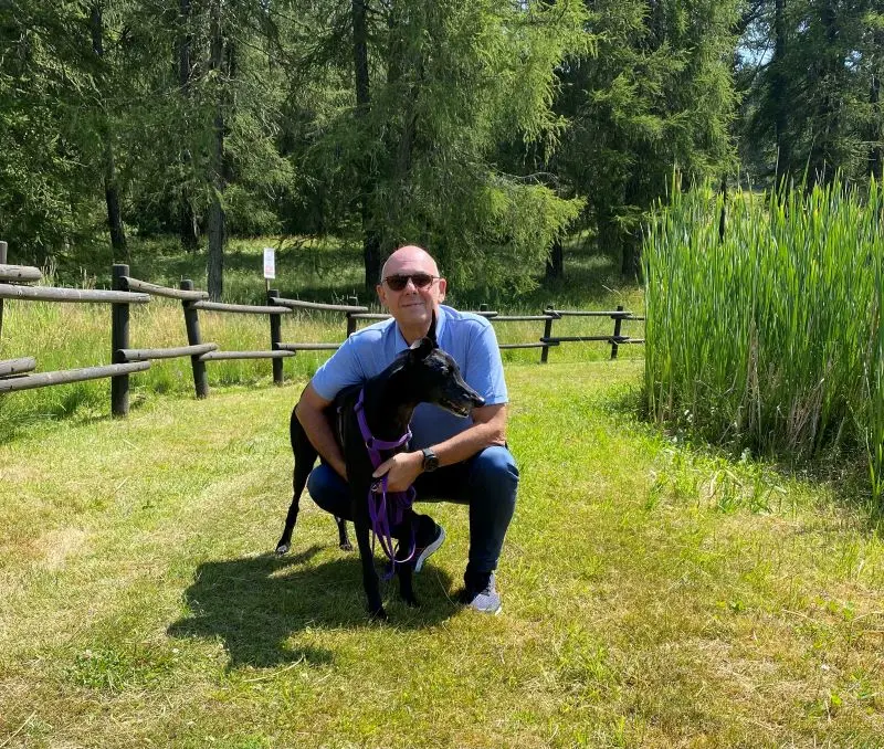 Lele con polo azzurra, abbraccia Lady, levriera nera con sullo sfondo una pineta