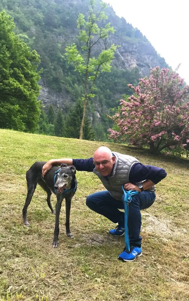 giovane uomo accosciato abbraccia la sua levriera nera, sullo sfondo la montagna