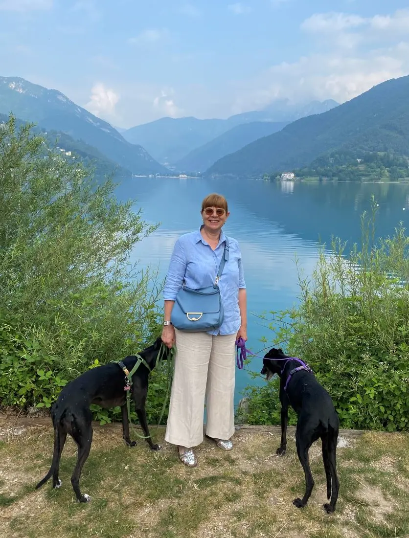 Elena con camicia azzurra e pantaloni bianchi, insieme alle sue levriere nere, con sullo sfondo il lago di Ledro e le montagne intorno.